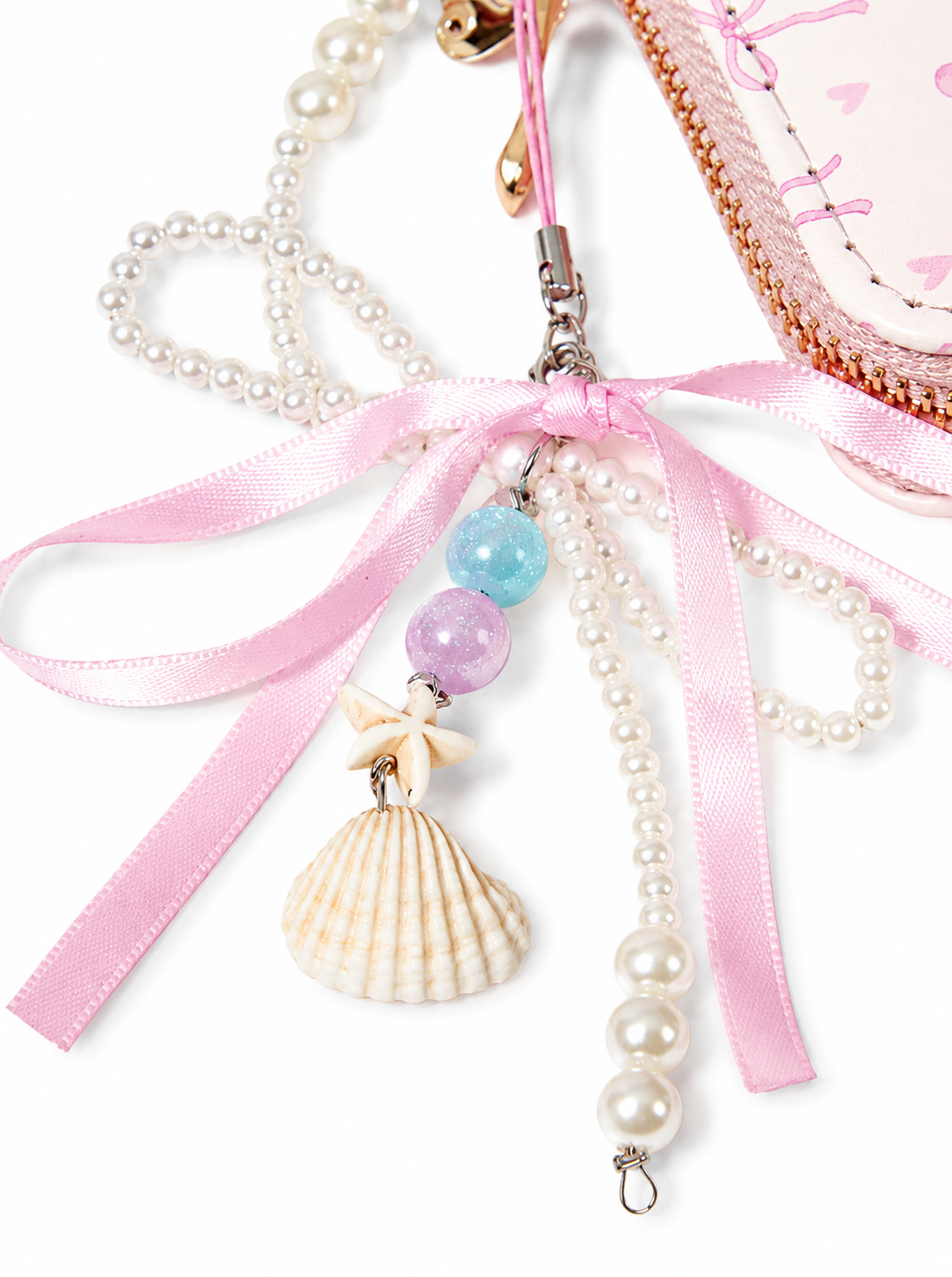 A close-up of a pastel pink and white decorative accessory with pearl strands, a pink satin ribbon, a seashell charm, pastel beads, and a small gold clasp, shown against a white background.